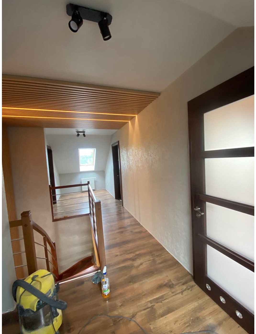 Korytarz po remoncie: brązowe panele, jasne ściany z efektem dekoracyjnym, drewniany sufit z oświetleniem LED. Widoczny odkurzacz i butelka po środku czyszczącym.