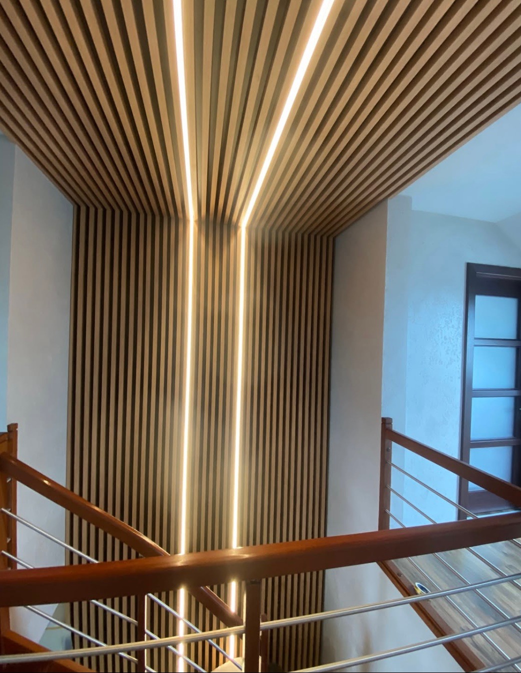 Drewniany sufit lamelowy z oświetleniem LED tworzący pionowe pasy światła. Widok z klatki schodowej, nowoczesny design.