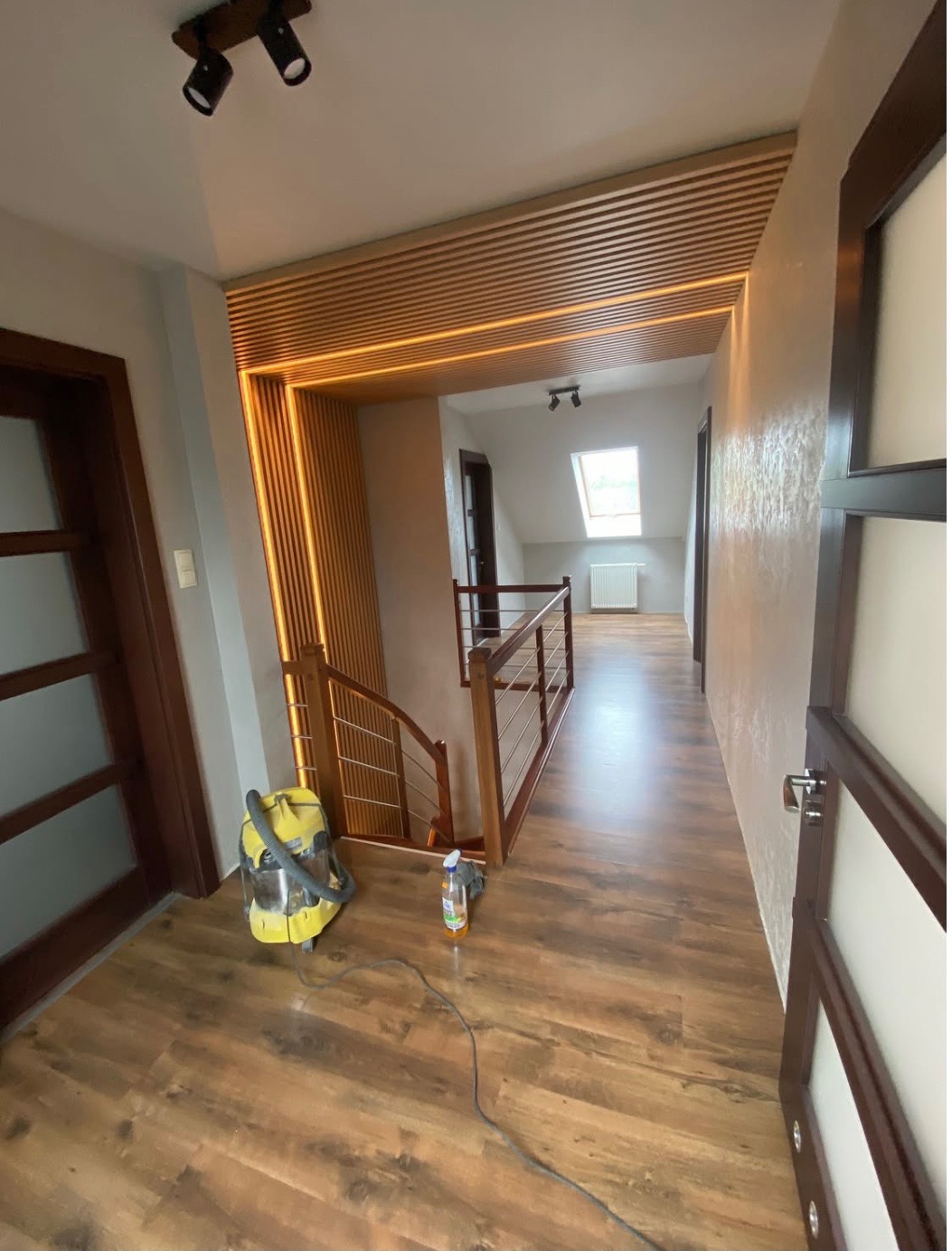 Wykończony korytarz z drewnianą podłogą, lamelami na suficie i podświetleniem LED. Widoczny odkurzacz i środek czyszczący, sugerujące prace porządkowe po remoncie.