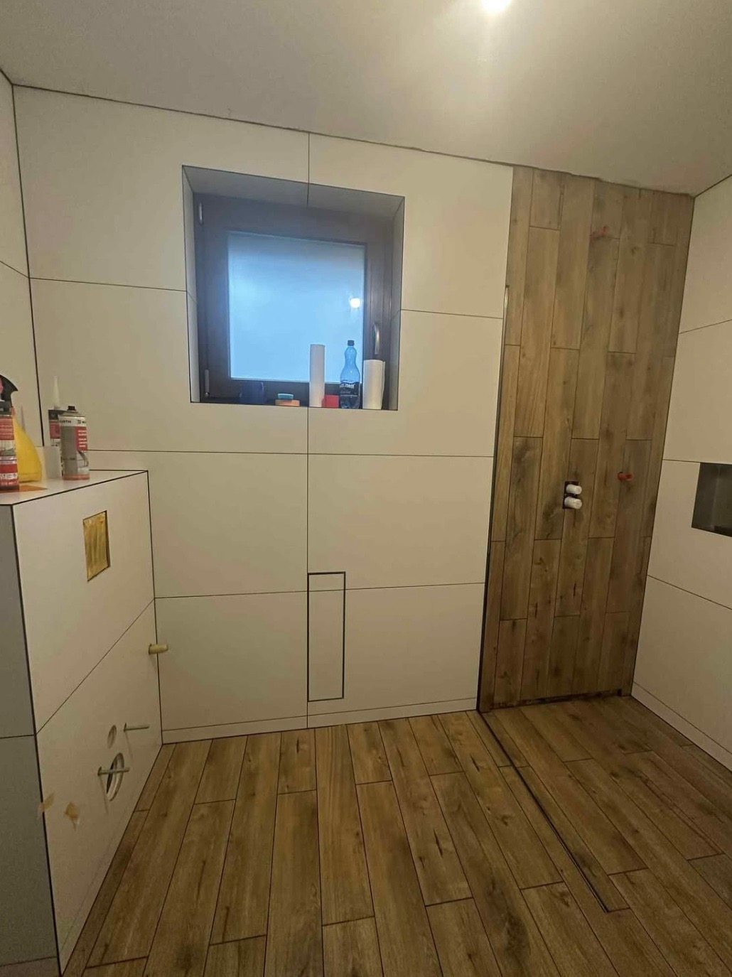 Wykończona łazienka z drewnopodobną podłogą i częściowo wykończonymi ścianami w płytkach. Widoczne okno i elementy instalacji.