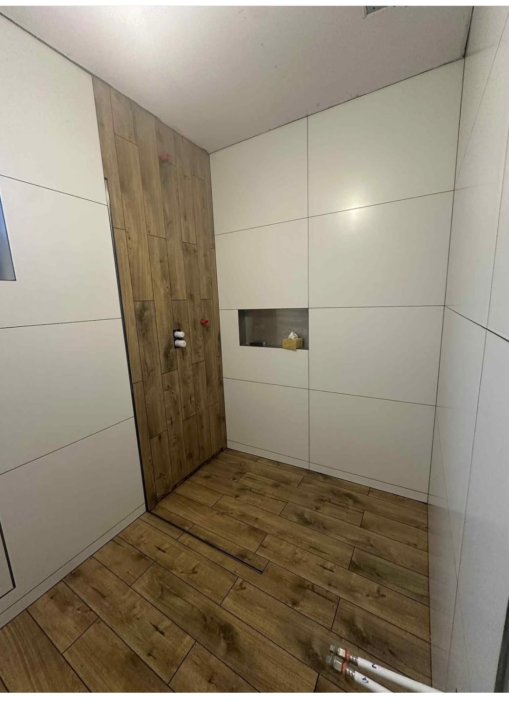 Nowoczesna łazienka z białą, kwadratową glazurą i akcentem drewna na ścianie prysznicowej oraz podłodze. Widoczne rury i wnęka w ścianie.