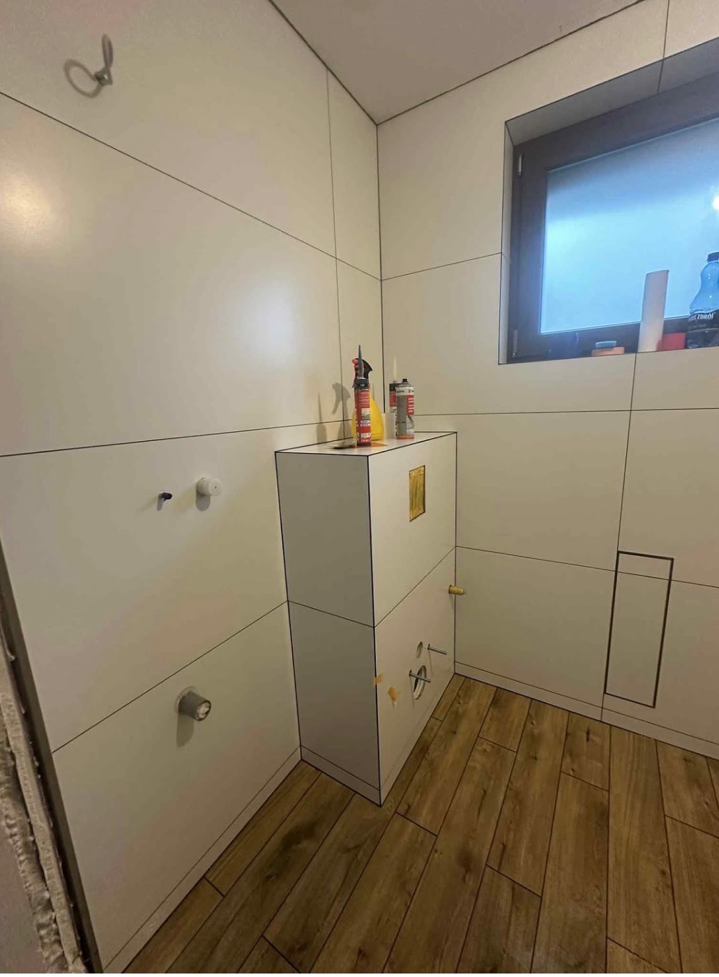 Łazienka w trakcie wykańczania: białe płytki z czarnym fugowaniem, drewnopodobna podłoga, zabudowa stelaża WC. Widoczne narzędzia i materiały budowlane.