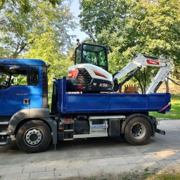 Mateusz Rudnik MATMEX - Niebieska ciężarówka MAN z białym minikoparką Bobcat E55z na platformie, zaparkowana na ulicy w słoneczny dzień.