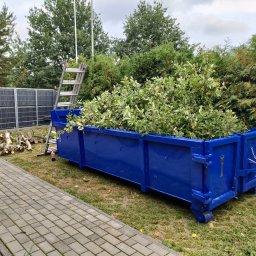 Mateusz Rudnik MATMEX - Niebieski kontener wypełniony obciętymi gałęziami i liśćmi stoi na trawniku obok ścieżki z kostki brukowej, w tle metalowe ogrodzenie i drabina oparta o drzewo.