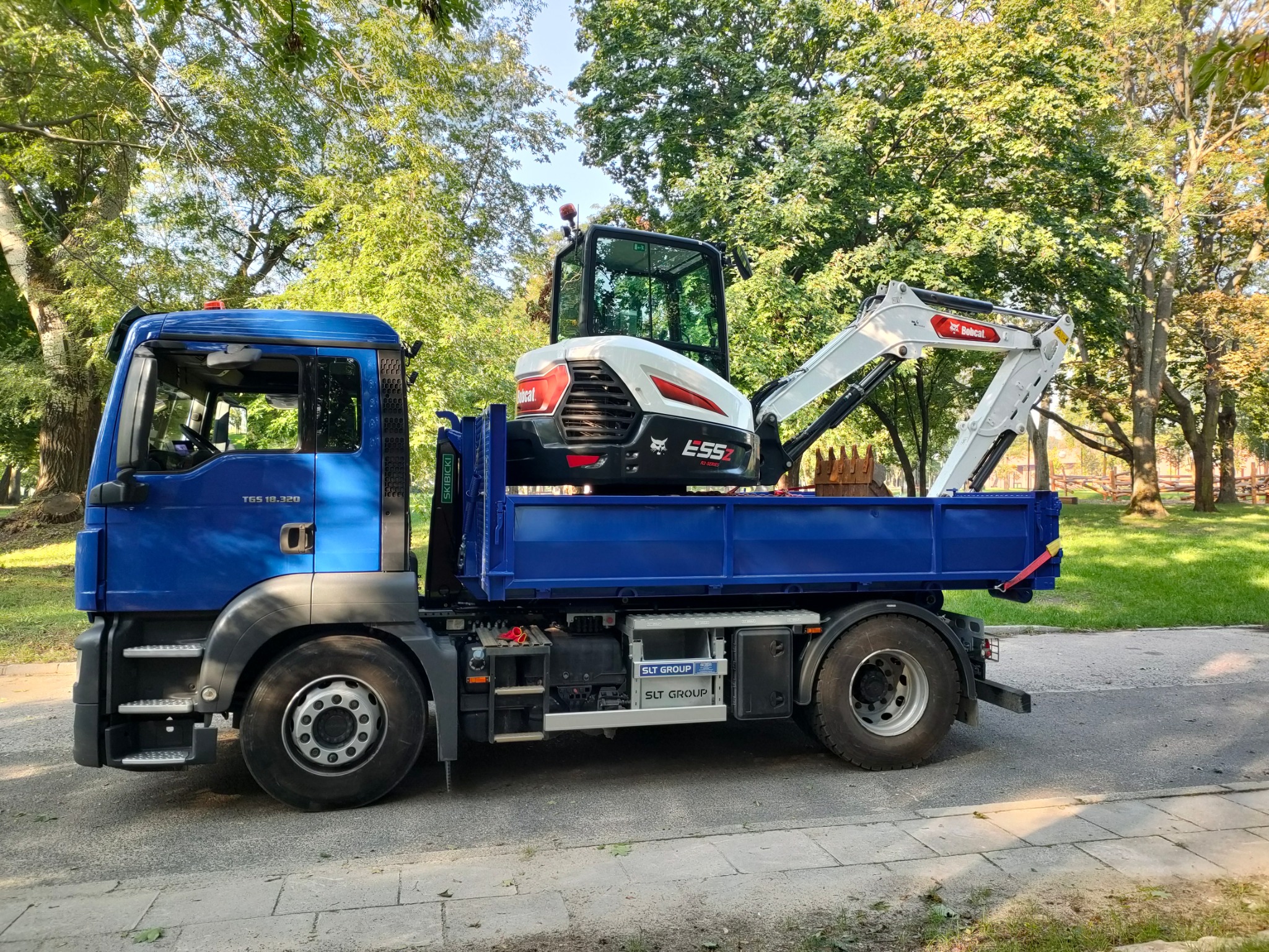 Niebieska ciężarówka MAN z białym minikoparką Bobcat E55z na platformie, zaparkowana na ulicy w słoneczny dzień.