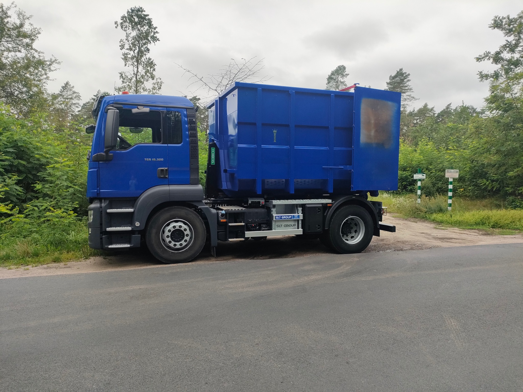 Niebieski samochód ciężarowy z kontenerem, zaparkowany na asfaltowej drodze, z widocznym fragmentem lasu w tle.