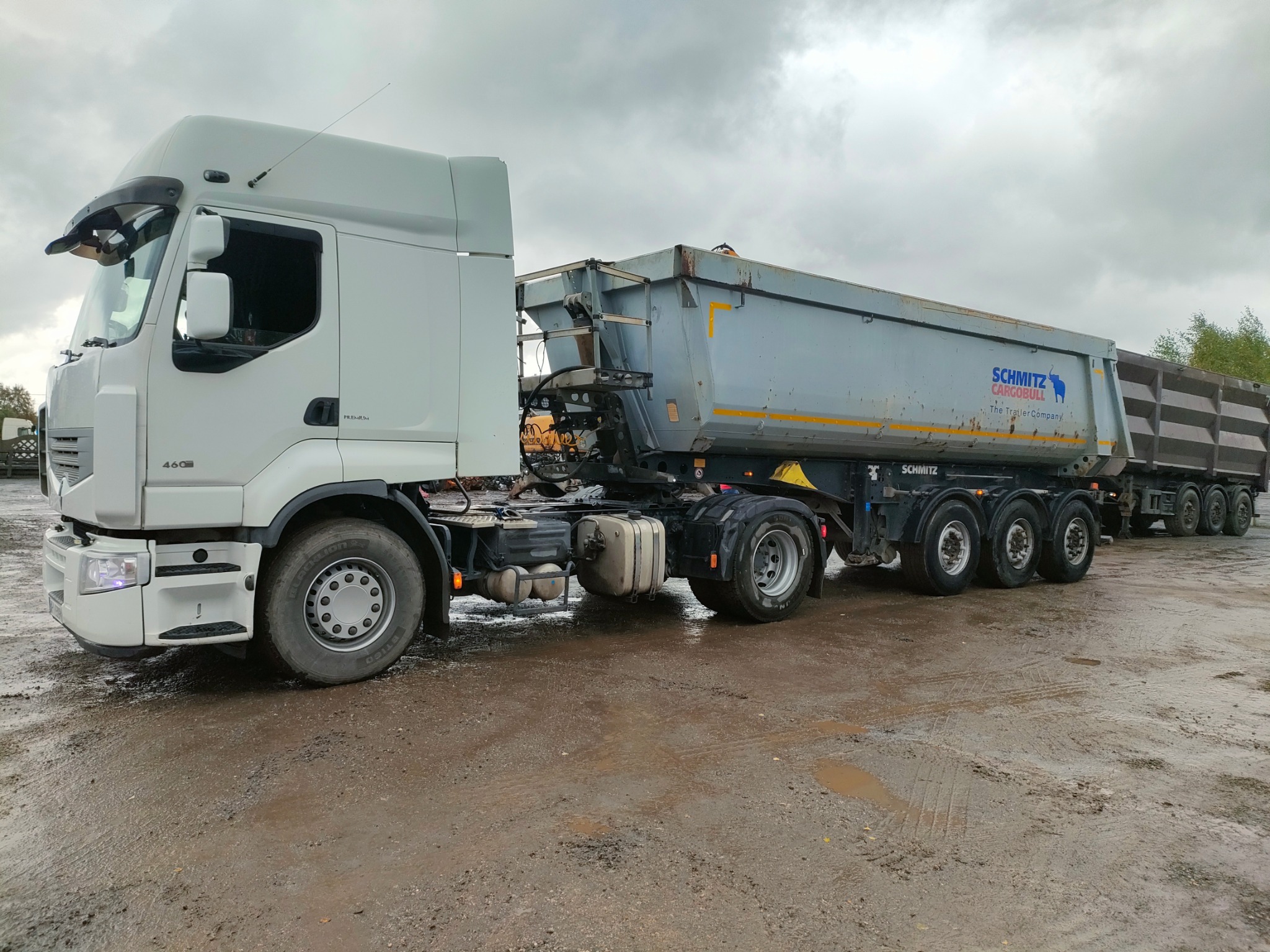 Ciężarówka Renault z naczepą Schmitz na błotnistym terenie. Widoczny fragment lasu w tle. Szary, pochmurny dzień. Perspektywa z boku, ukazująca detale pojazdu.