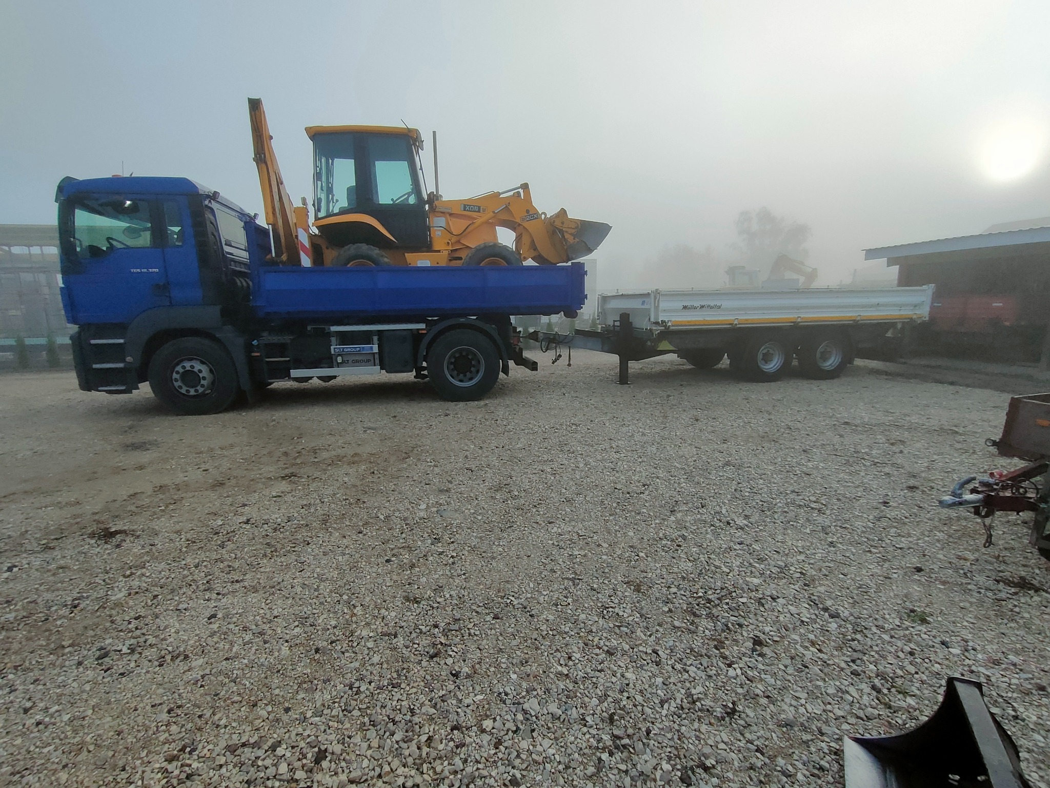 Niebieska ciężarówka z żółtą koparką na platformie i przyczepą w tle, w warunkach mglistej pogody. Widoczny żwir na ziemi.