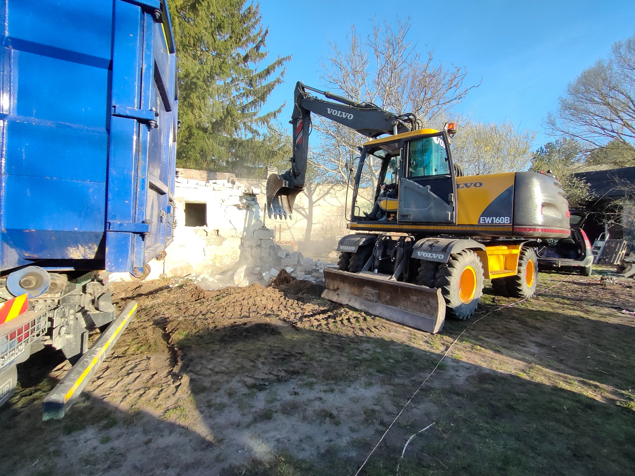 Żółta koparka Volvo EW160B burzy fragment murowanej ściany, obok stoi niebieski kontener na gruz. Praca w toku, ziemia wokół rozjeżdżona.
