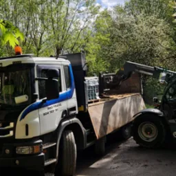 Ciężarówka Scania z kontenerem i miniładowarką na drodze, widoczne drzewa w tle, częściowo ocienione ujęcie.