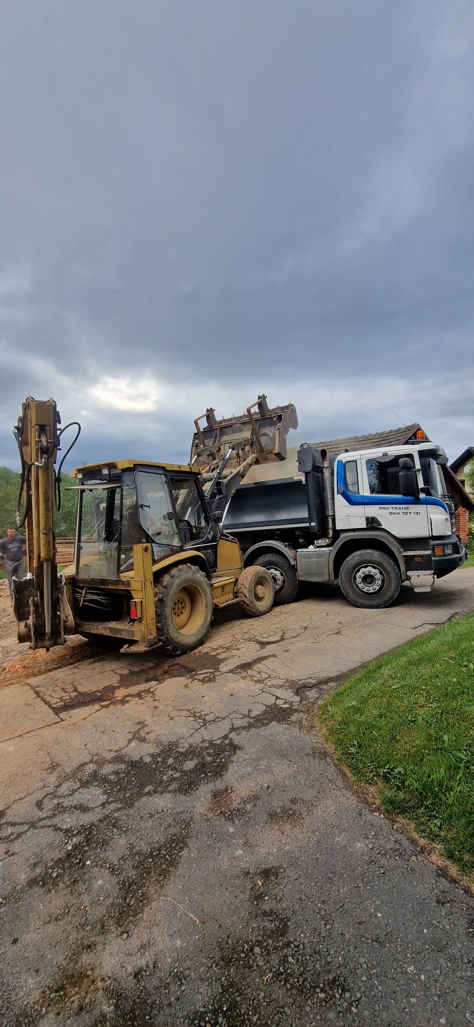 Żółta koparko-ładowarka i ciężarówka z wywrotką podczas załadunku na podmokłym terenie, widoczne logo firmy transportowej na kabinie ciężarówki.