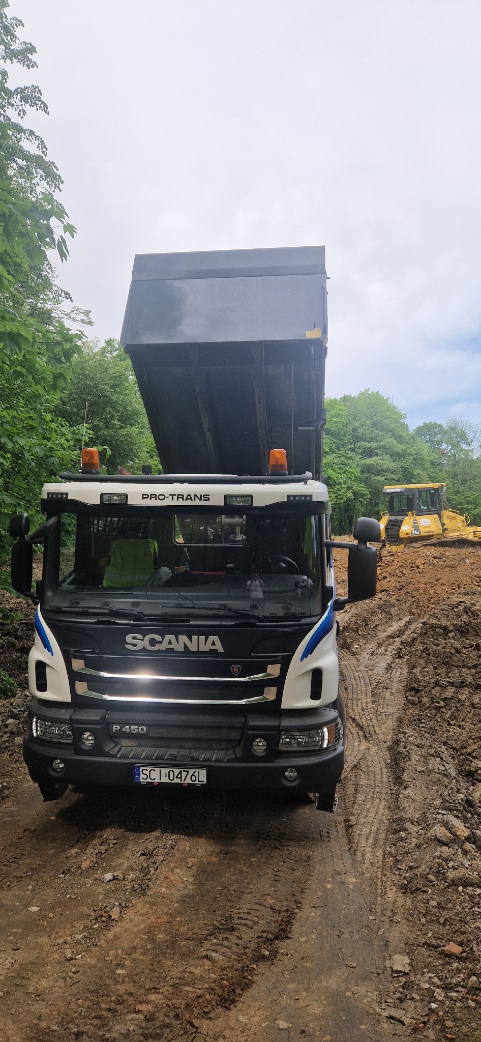 Ciężarówka Scania P450 z podniesioną skrzynią ładunkową na błotnistej drodze, w tle widoczna żółta spycharka, otoczenie leśne.