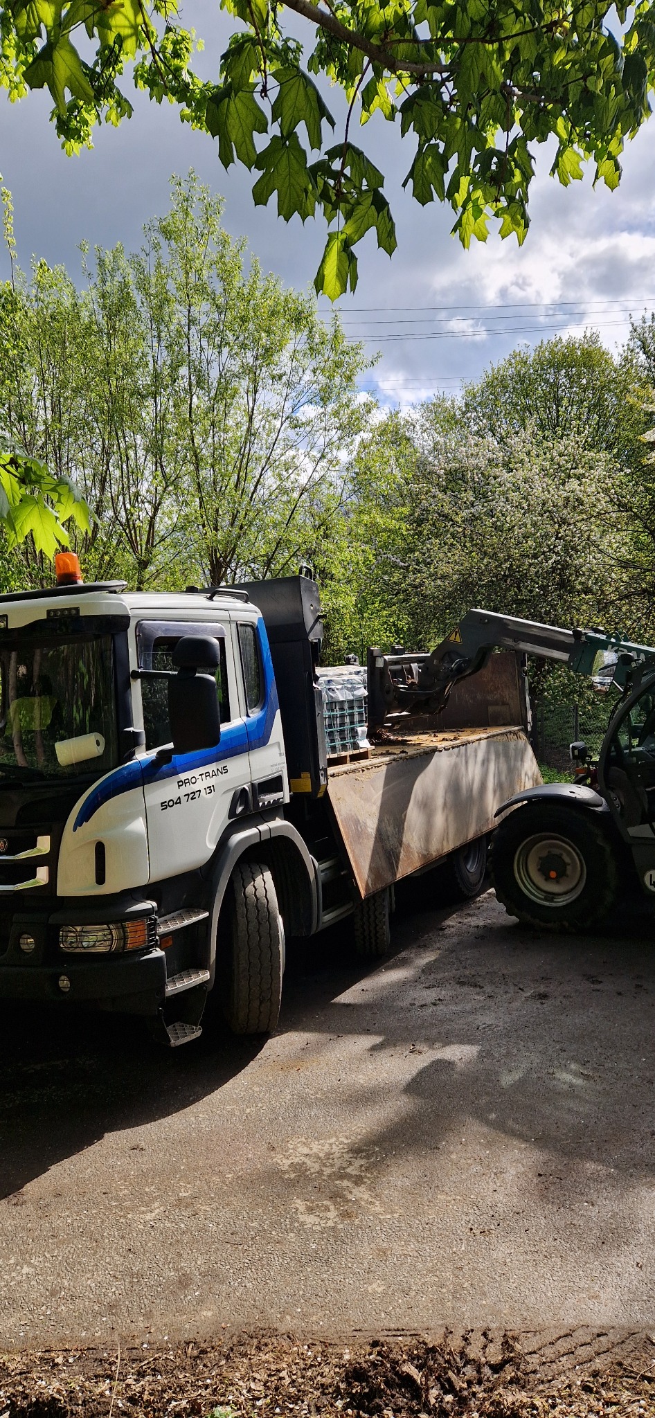 Ciężarówka Scania z kontenerem i miniładowarką na drodze, widoczne drzewa w tle, częściowo ocienione ujęcie.