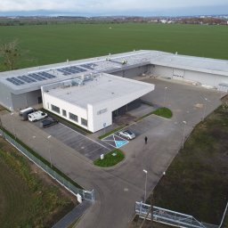 EXIGOBUD RAFAŁ STEFAŃSKI - Widok z lotu ptaka na budynek usługowo-handlowy z panelami słonecznymi na dachu, parkingiem z miejscem dla niepełnosprawnych i zagospodarowanym terenem zielonym.