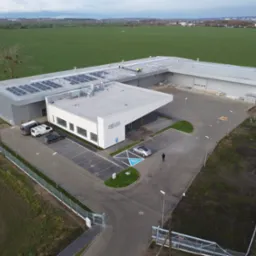 Widok z lotu ptaka na budynek usługowo-handlowy z panelami słonecznymi na dachu, parkingiem z miejscem dla niepełnosprawnych i zagospodarowanym terenem zielonym.