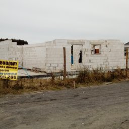 EXIGOBUD RAFAŁ STEFAŃSKI - Budowa domu jednorodzinnego w technologii tradycyjnej: surowe ściany z betonu komórkowego, widoczny baner z napisem 'KIEROWNIK BUDOWY' i numerem telefonu, w tle inny dom i drzewa.