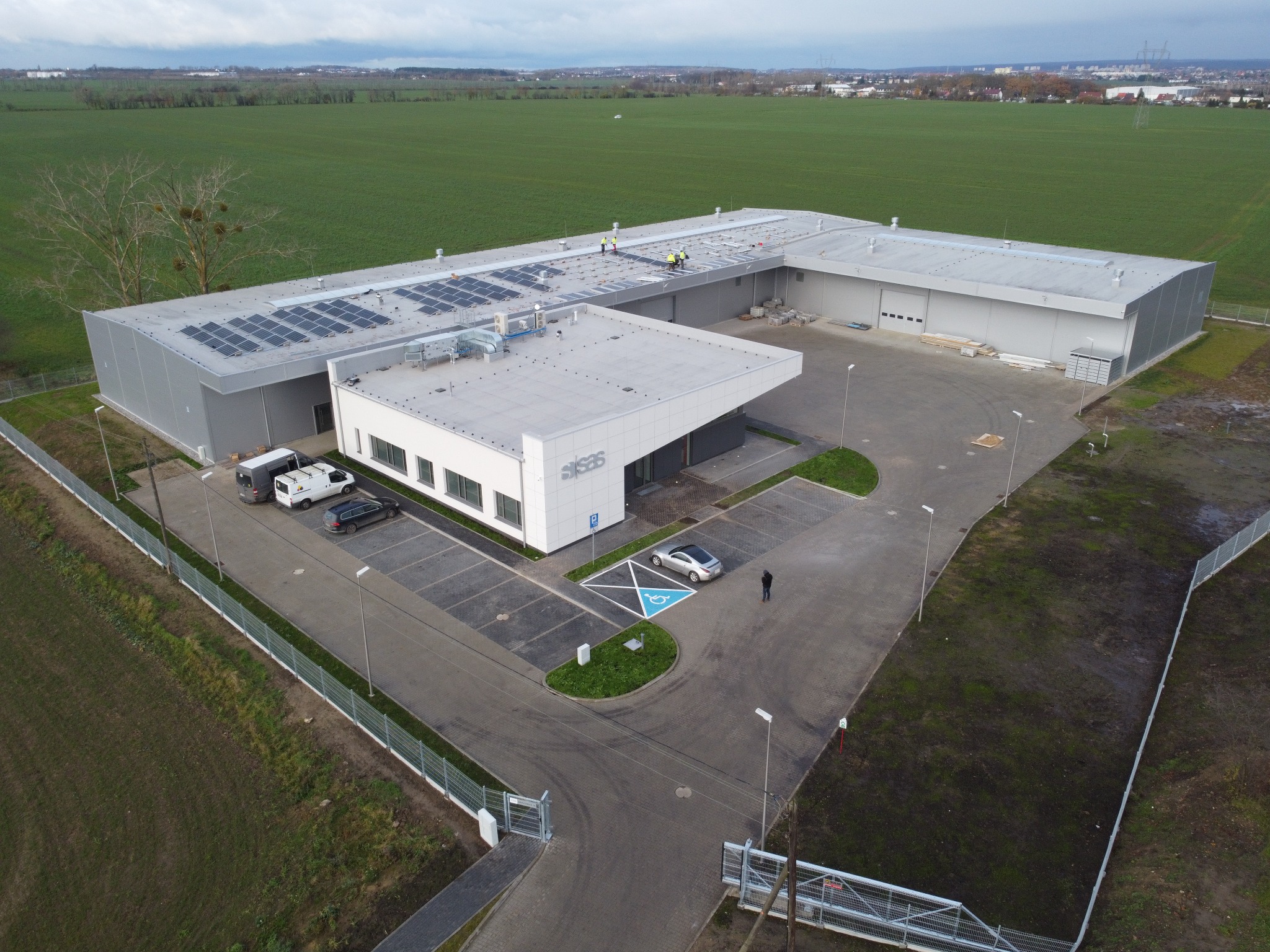 Widok z lotu ptaka na budynek usługowo-handlowy z panelami słonecznymi na dachu, parkingiem z miejscem dla niepełnosprawnych i zagospodarowanym terenem zielonym.