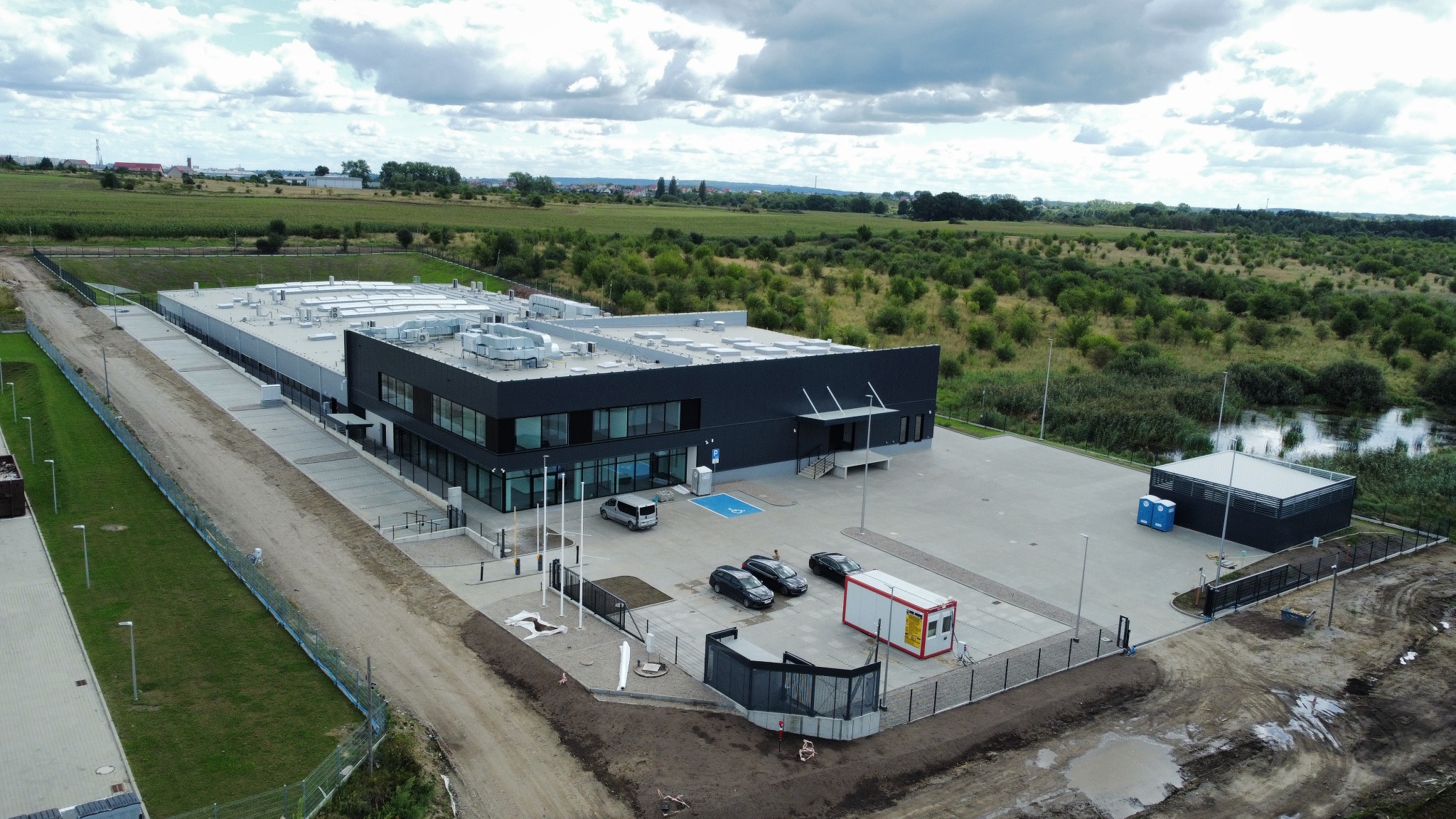Zakończona budowa hali produkcyjno-magazynowej z częścią biurową i zagospodarowaniem terenu, widok z lotu ptaka na budynek z płaskim dachem, parkingiem z samochodami osobowymi, kontenerem biurowym...