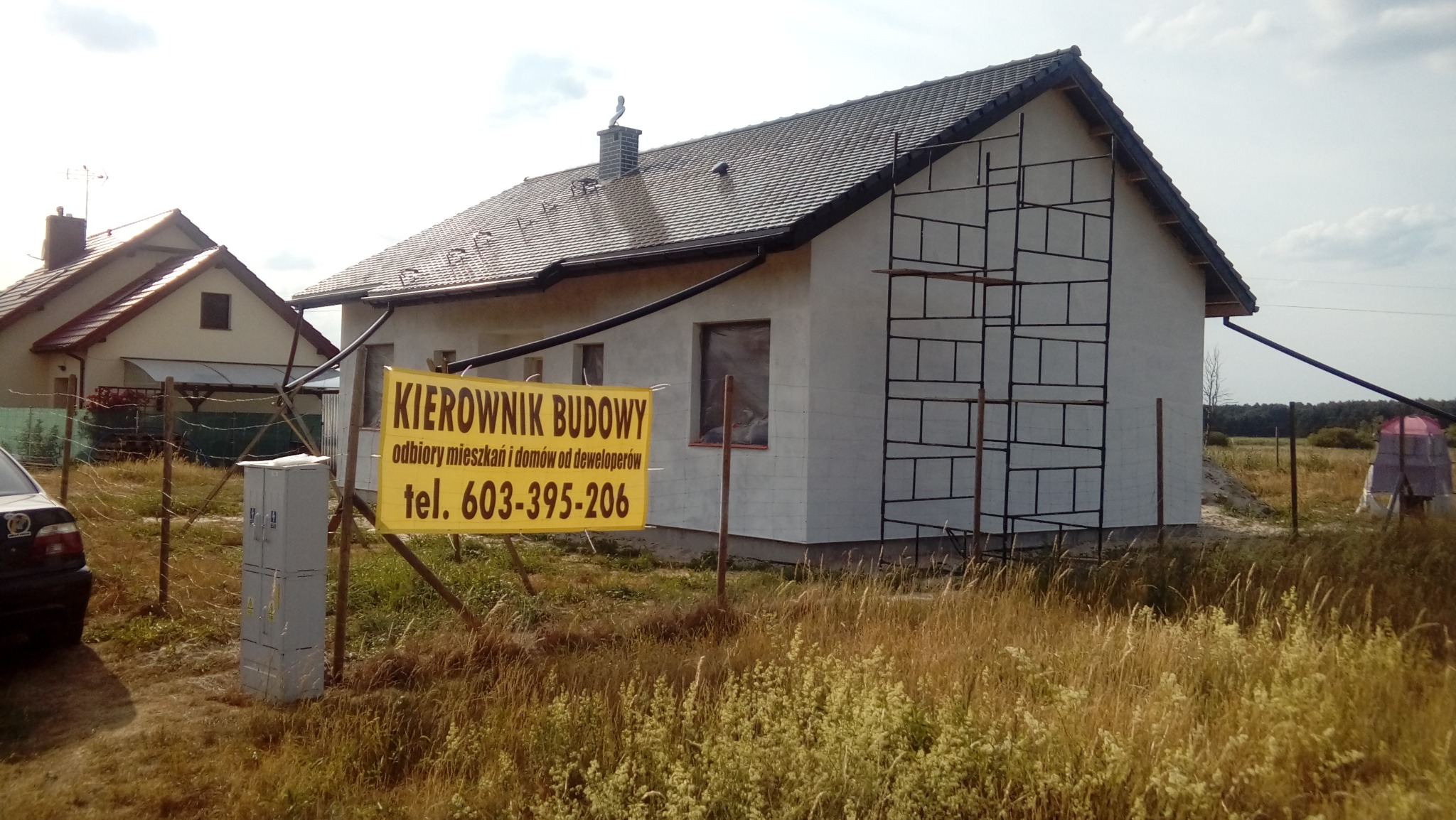 Dom jednorodzinny w trakcie budowy, z zamontowanym rusztowaniem przy ścianie szczytowej, widoczny baner informacyjny z numerem telefonu.