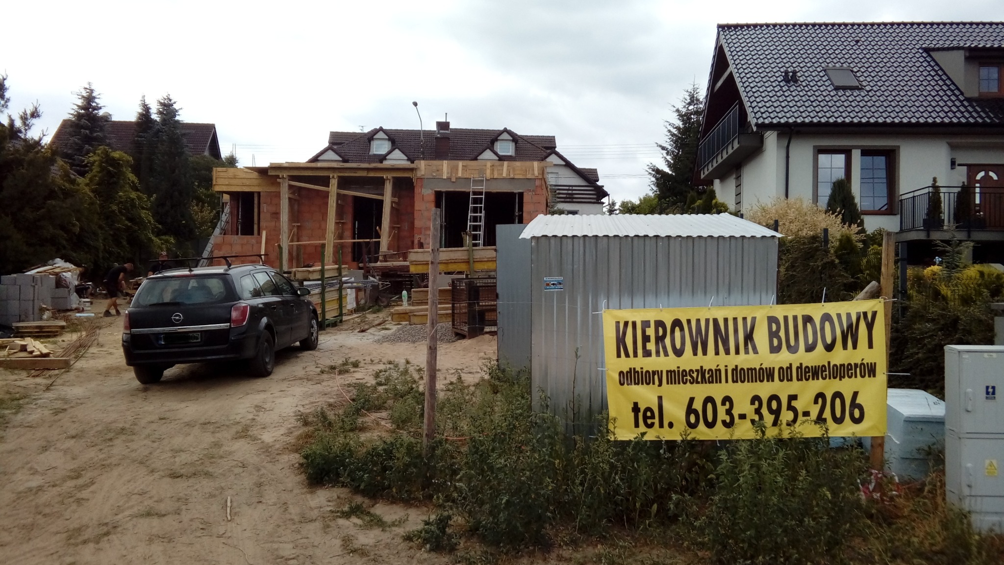 Budowa domu jednorodzinnego w trakcie realizacji, widoczna konstrukcja z czerwonej cegły, drewniane elementy szalunku, drabina oparta o ścianę, na pierwszym planie baner informacyjny z napisem...