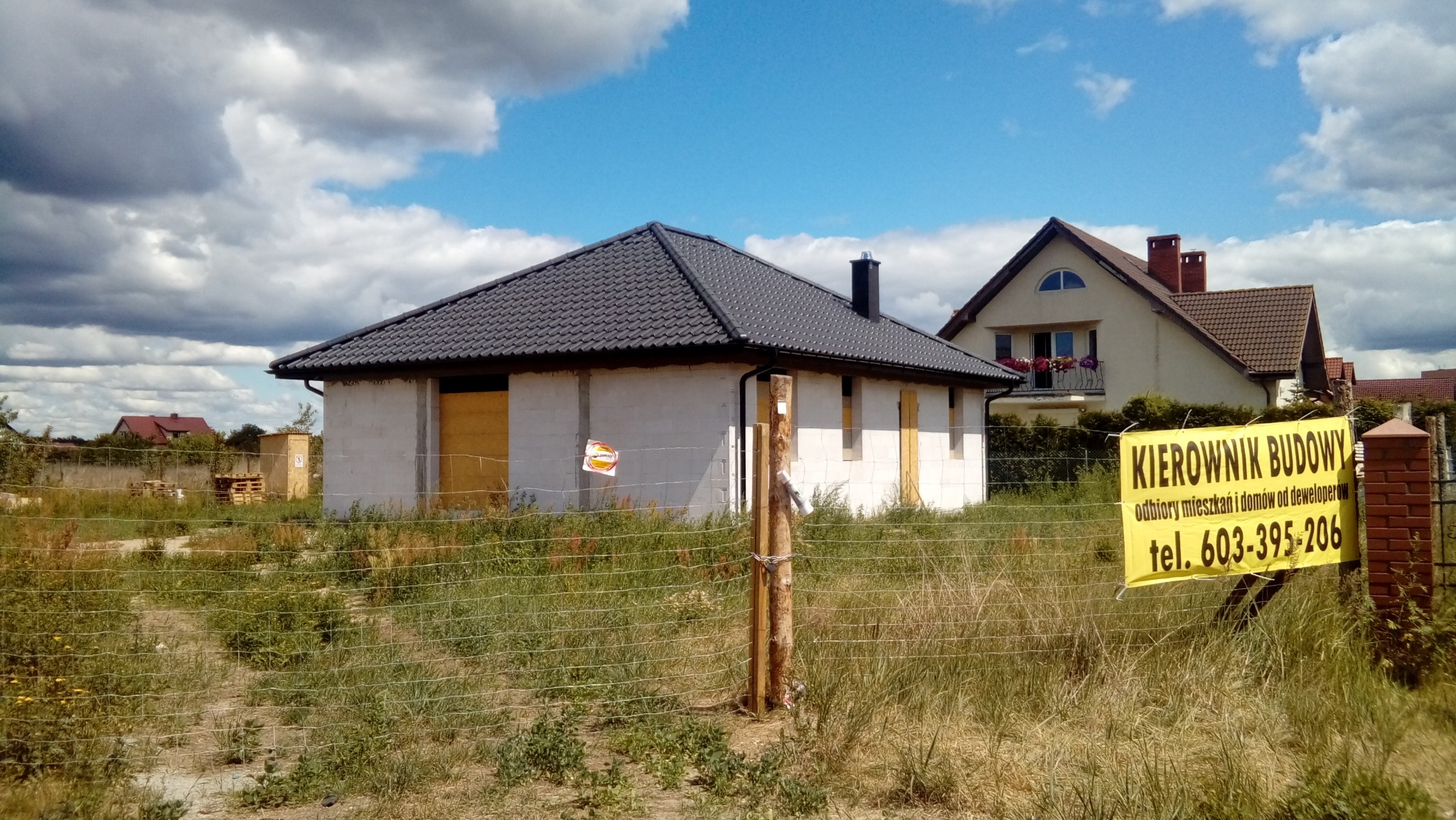 Niedokończony dom jednorodzinny z szarym dachem i zamurowanymi oknami, widoczny przez siatkę ogrodzeniową z żółtym banerem reklamowym z napisem i numerem telefonu, w tle drugi dom z balkonem...