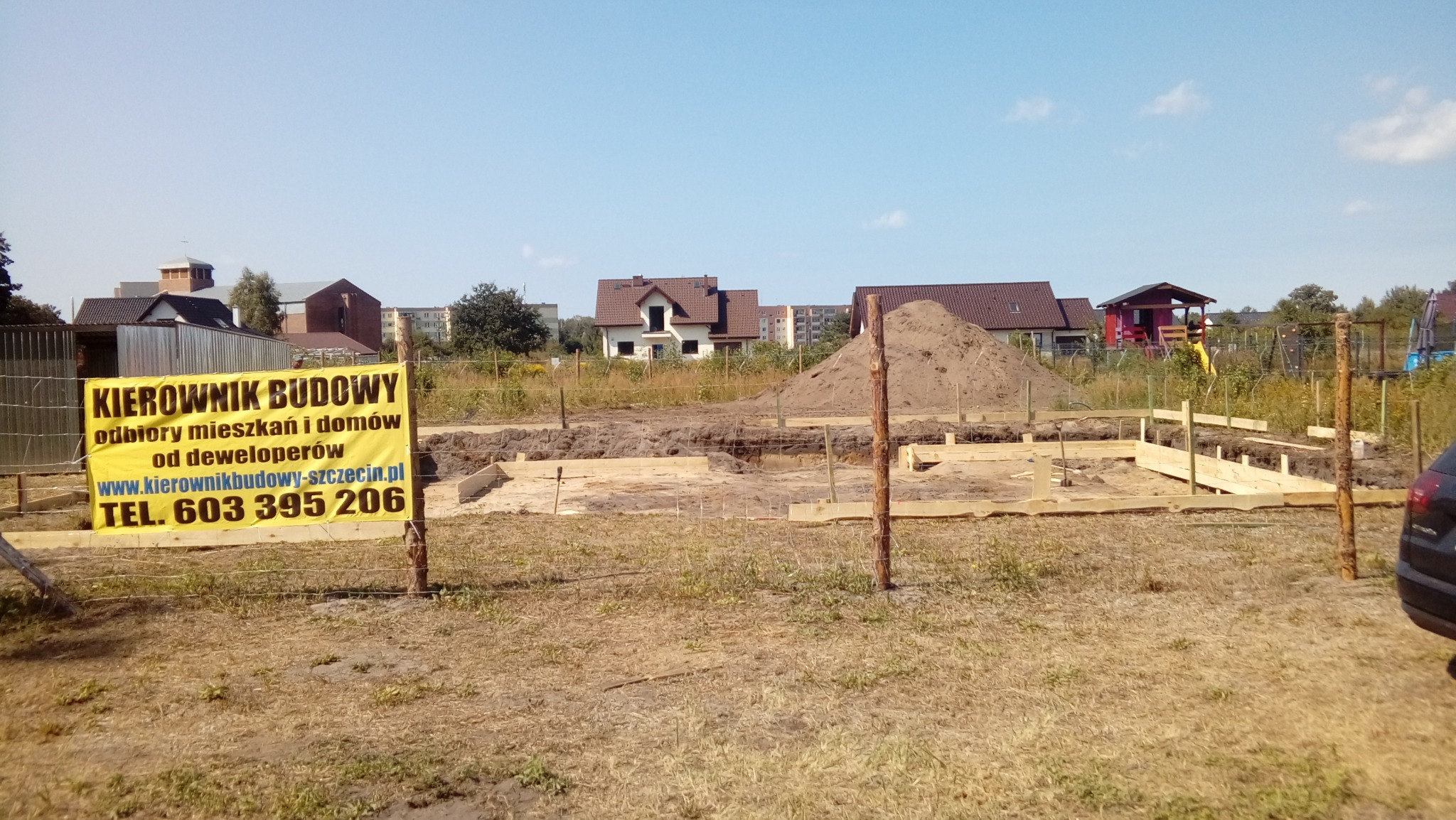 Fundamenty domu jednorodzinnego w trakcie budowy, widoczna żółta tablica informacyjna z napisem 'Kierownik Budowy', w tle zabudowa jednorodzinna i wielorodzinna.