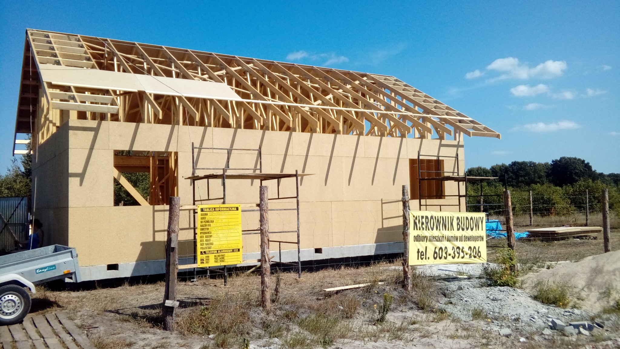 Budowa domu jednorodzinnego w technologii drewnianej w trakcie realizacji, widoczne elementy konstrukcyjne dachu, ściany z płyt OSB, rusztowania oraz tablica informacyjna budowy i baner z numerem...