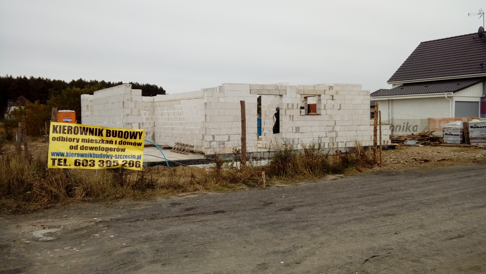 Budowa domu jednorodzinnego w technologii tradycyjnej: surowe ściany z betonu komórkowego, widoczny baner z napisem 'KIEROWNIK BUDOWY' i numerem telefonu, w tle inny dom i drzewa.