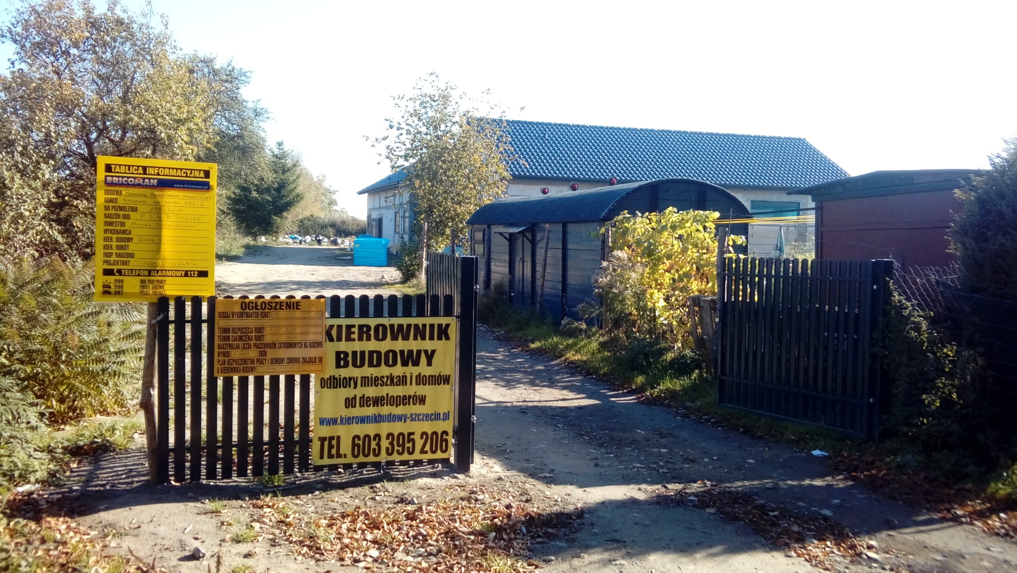 Teren budowy z tablicami informacyjnymi i ogłoszeniem o kierowniku budowy, widoczne budynki w tle.