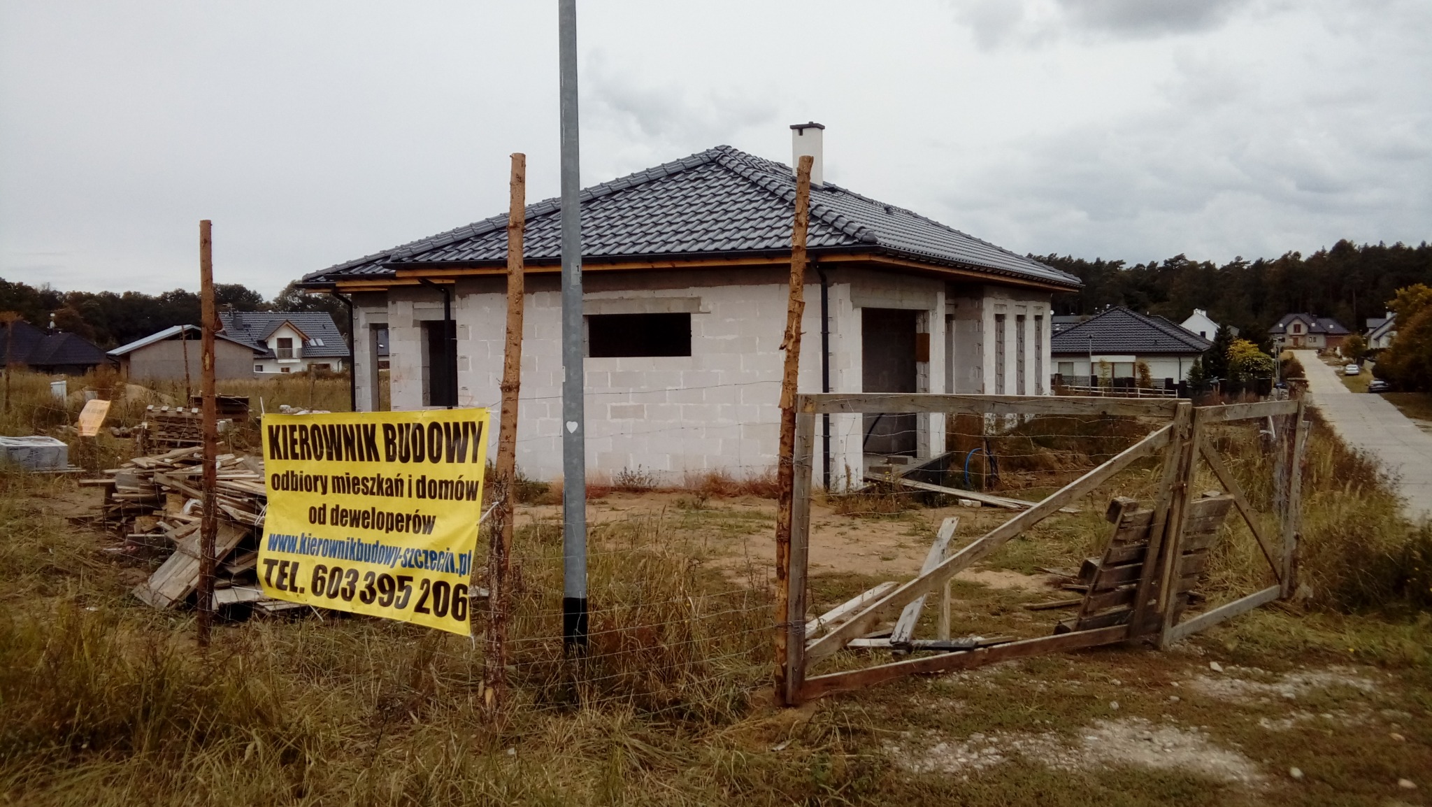 Surowy stan domu jednorodzinnego z szarą dachówką, widoczny przez prowizoryczne ogrodzenie i żółty baner z napisem 'KIEROWNIK BUDOWY' oraz numerem telefonu, z pochmurnym niebem w tle.