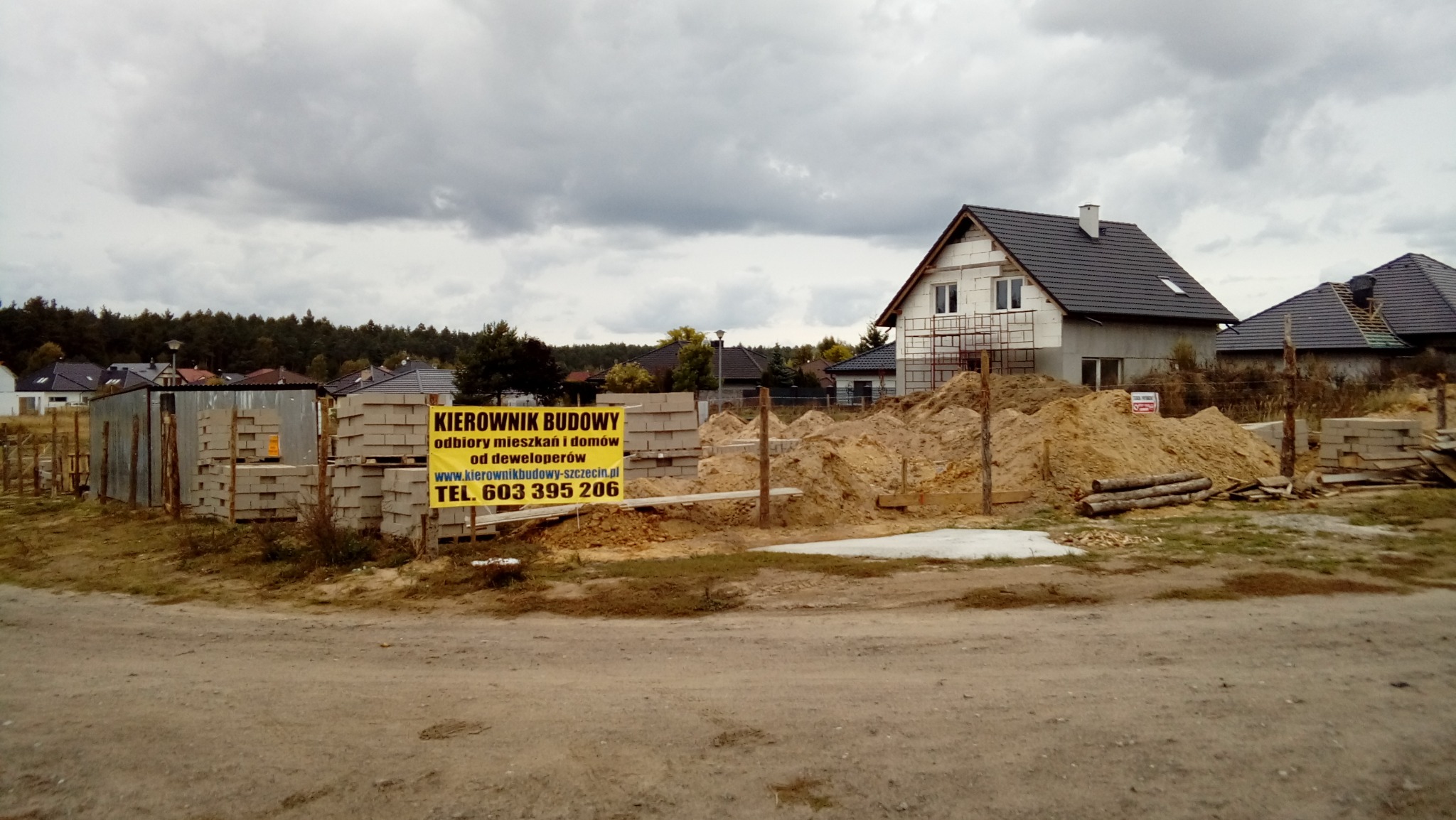 Budowa domu jednorodzinnego w stanie surowym otwartym, widoczne bloczki betonowe, piasek, oraz żółty baner reklamowy z napisem 'KIEROWNIK BUDOWY' i numerem telefonu, pochmurne niebo w tle.