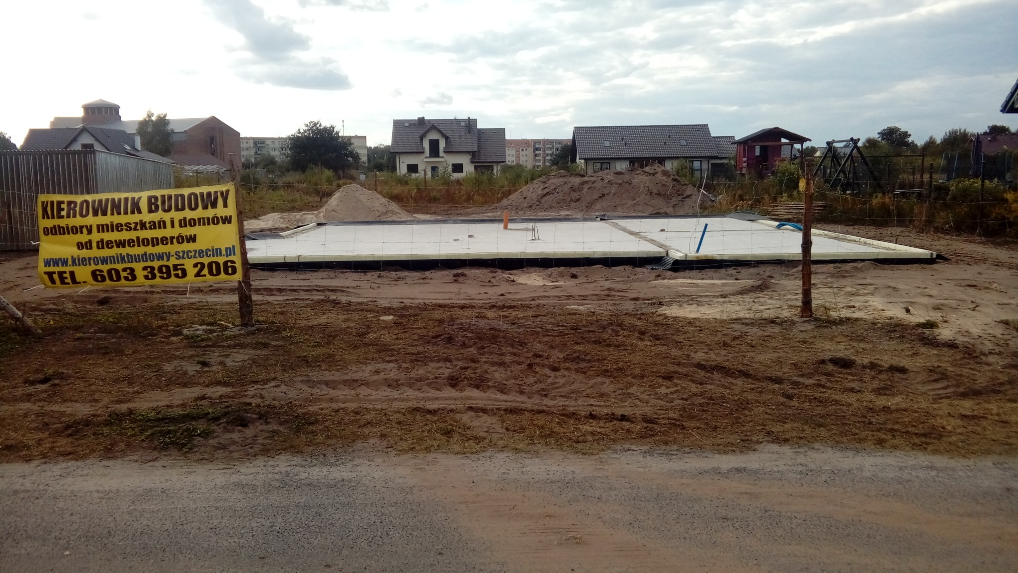 Przygotowanie fundamentów pod budowę domu jednorodzinnego: wylana płyta fundamentowa z izolacją termiczną, widoczny żółty baner z napisem 'KIEROWNIK BUDOWY' i numerem telefonu, w tle zabudowa...