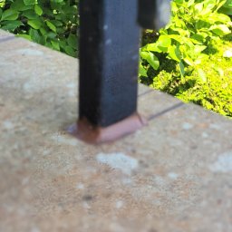 Cts fiszka - Fragment balustrady balkonowej z czarną metalową poręczą, zamocowaną do jasnobrązowej płytki, z zielenią w tle, sfotografowane z bliska.