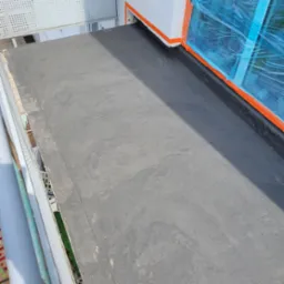 Wykończona, czarna, bitumiczna izolacja balkonu z widocznymi detalami uszczelnienia przy ścianie i zabezpieczonym oknem folią malarską.