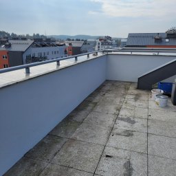 Cts fiszka - Balkon na dachu budynku wielorodzinnego z widokiem na zabudowę miejską. Świeżo pomalowana ściana balkonu, brudne płytki podłogowe i puszki z farbą w rogu sugerują prace wykończeniowe.