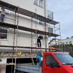 Cts fiszka - Trzech pracowników na rusztowaniu przy elewacji budynku wielorodzinnego, jeden stoi w skrzyni czerwonego samochodu dostawczego, widoczna tabliczka ostrzegawcza 'Uwaga! Roboty na wysokości'.