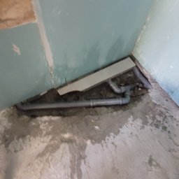 Cts fiszka - Niedokończona łazienka z odsłoniętymi rurami kanalizacyjnymi w rogu, ściany pokryte wstępnie nałożoną gładzią, widoczne ślady po szpachlowaniu.