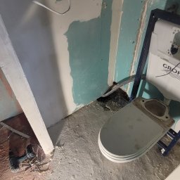 Cts fiszka - Niedokończona łazienka w trakcie remontu: widoczne fragmenty ścian z niepełnymi gładziami, stelaż podtynkowy WC Grohe, narzędzia i pozostałości po pracach na podłodze, niezakryte przewody elektryczne.