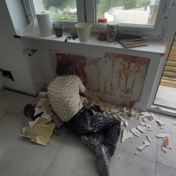 Cts fiszka - Mężczyzna w koszuli w kratę, w ubrudzonych farbą spodniach, skuwa starą warstwę tynku ze ściany pod oknem, widoczne liczne odłamki i resztki materiału na podłodze.