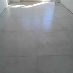 Ułożona podłoga z dużych, matowych, szarych płytek w nowym, jasnym pomieszczeniu, z widocznym fragmentem ściany z otworem na instalację elektryczną i fragmentem krzesła przez otwarte drzwi.