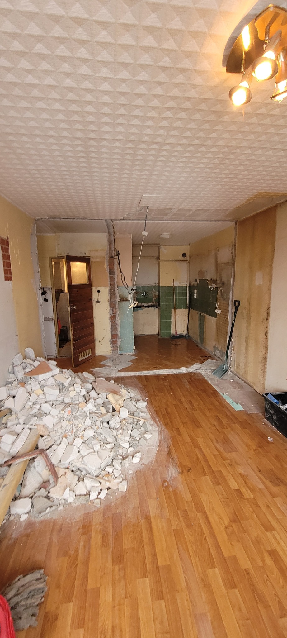 Demontaż ściany działowej w trakcie remontu mieszkania, widoczne odsłonięte instalacje elektryczne, sterta gruzu na podłodze, fragmenty starej glazury w kuchni w głębi.