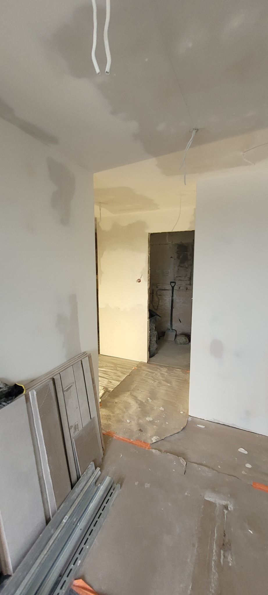 Wnętrze w trakcie remontu: przygotowane płyty kartonowo-gipsowe o szarym odcieniu opierają się o ścianę, widoczne profile metalowe na podłodze, w tle otwór drzwiowy z widoczną łopatą...