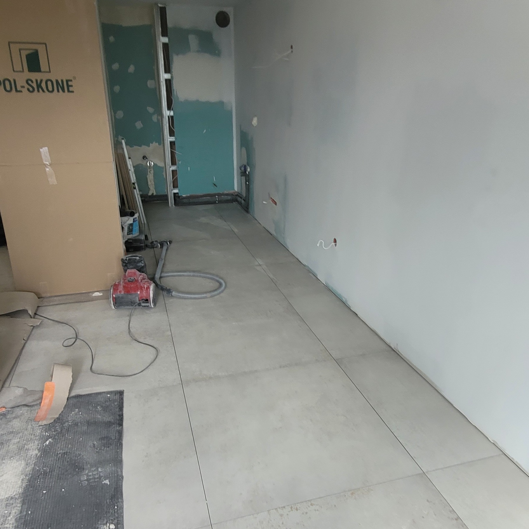 Wnętrze w trakcie remontu z ułożonymi jasnymi płytkami podłogowymi, widoczny karton z napisem POL-SKONE, odkurzacz przemysłowy i fragment ściany w trakcie przygotowywania pod gładzie.
