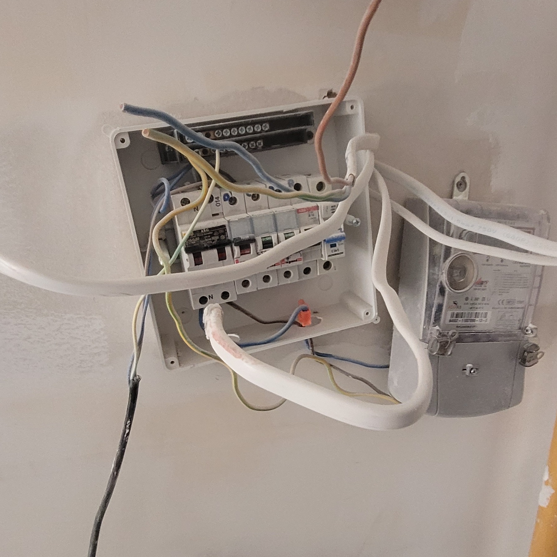 Rozdzielnica elektryczna z bezpiecznikami i licznikiem, widoczne przewody w różnych kolorach, w trakcie instalacji na ścianie.