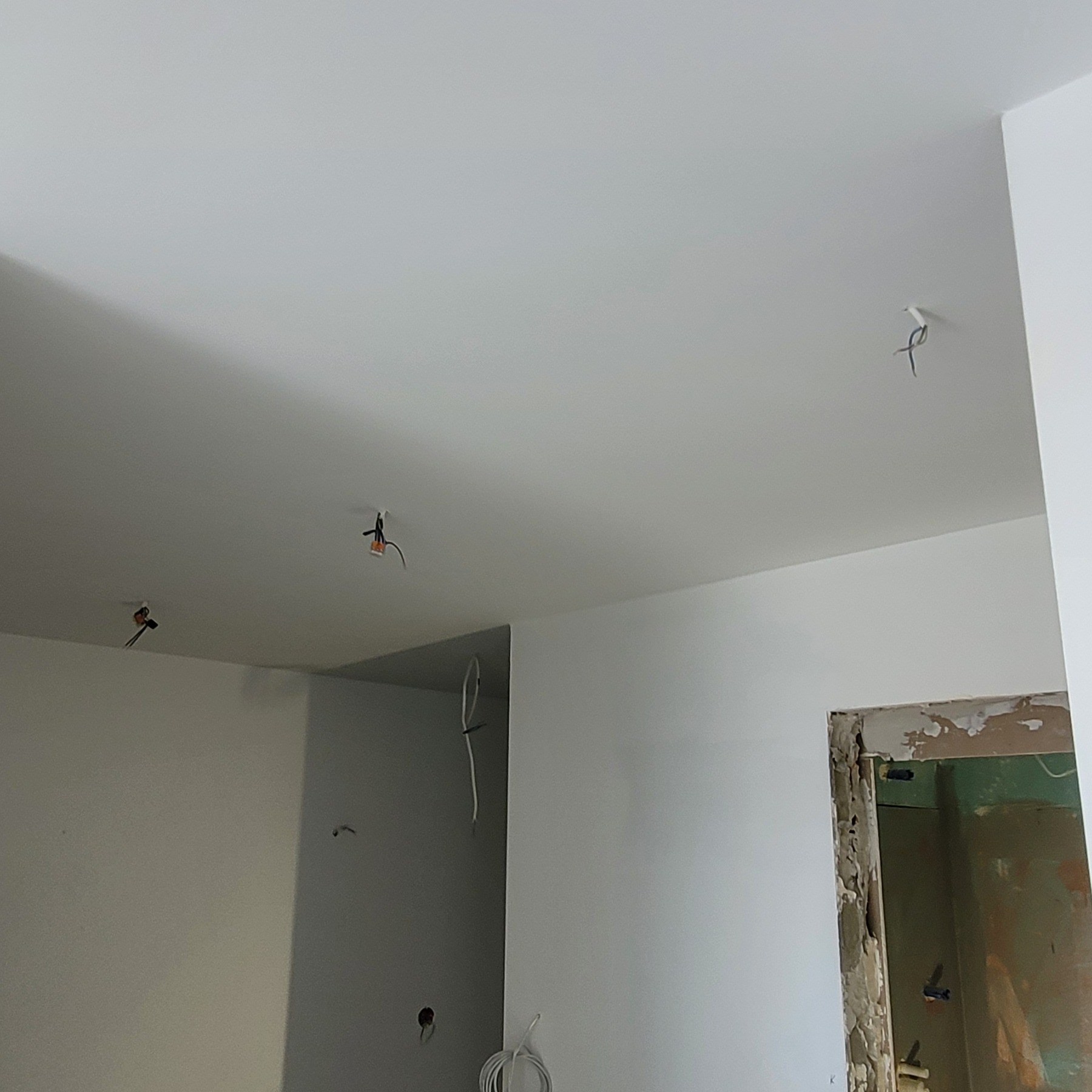 Biały, świeżo pomalowany sufit z wystającymi przewodami elektrycznymi, przygotowany do montażu oświetlenia, z widocznym fragmentem ściany i otworem drzwiowym w tle.