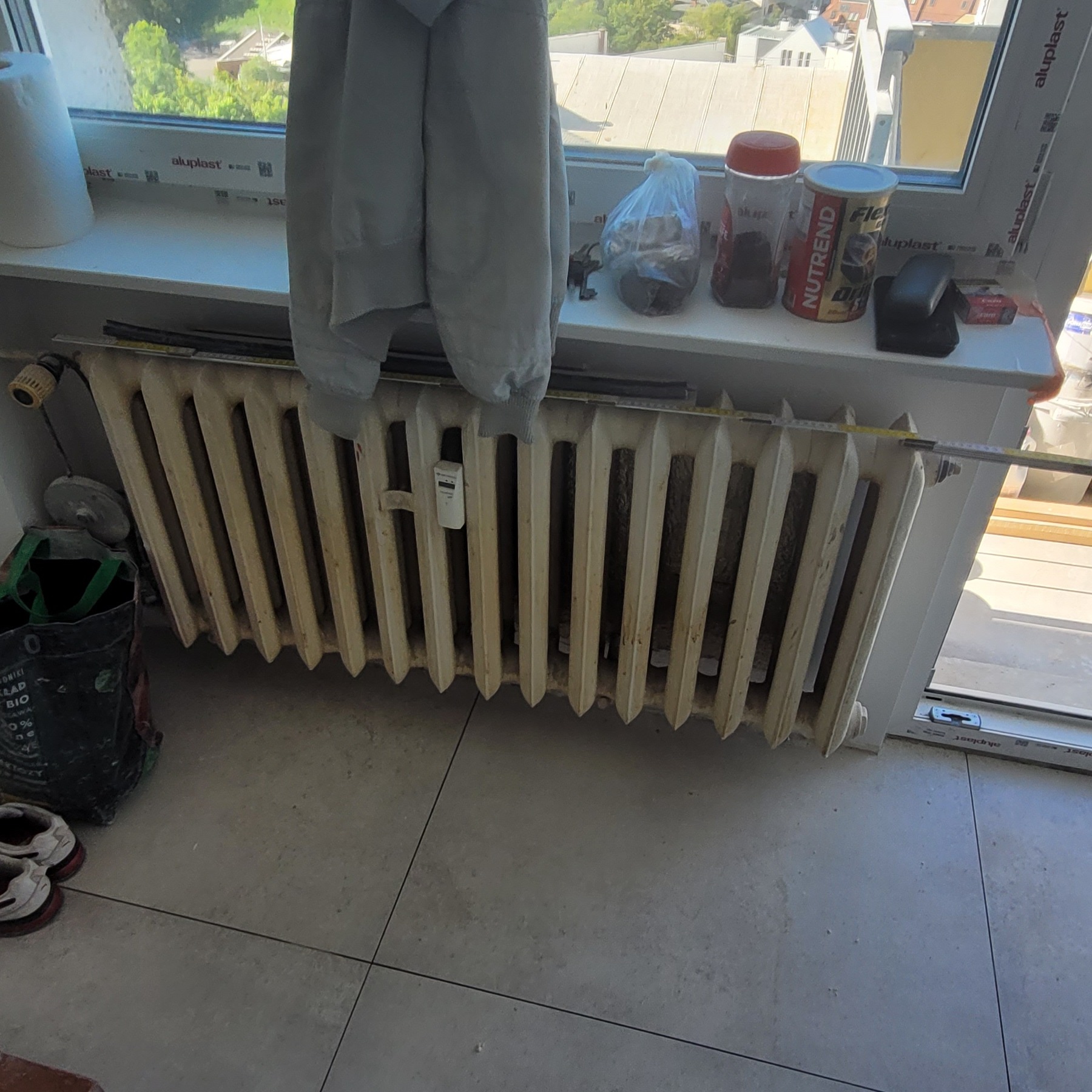 Stary, żeliwny grzejnik żeberkowy z termostatem, stojący przy oknie PCV, obok torba na śmieci, para butów i wisząca bluza. Na parapecie leżą różne przedmioty, w tym miarka i opakowania po odżywkach.