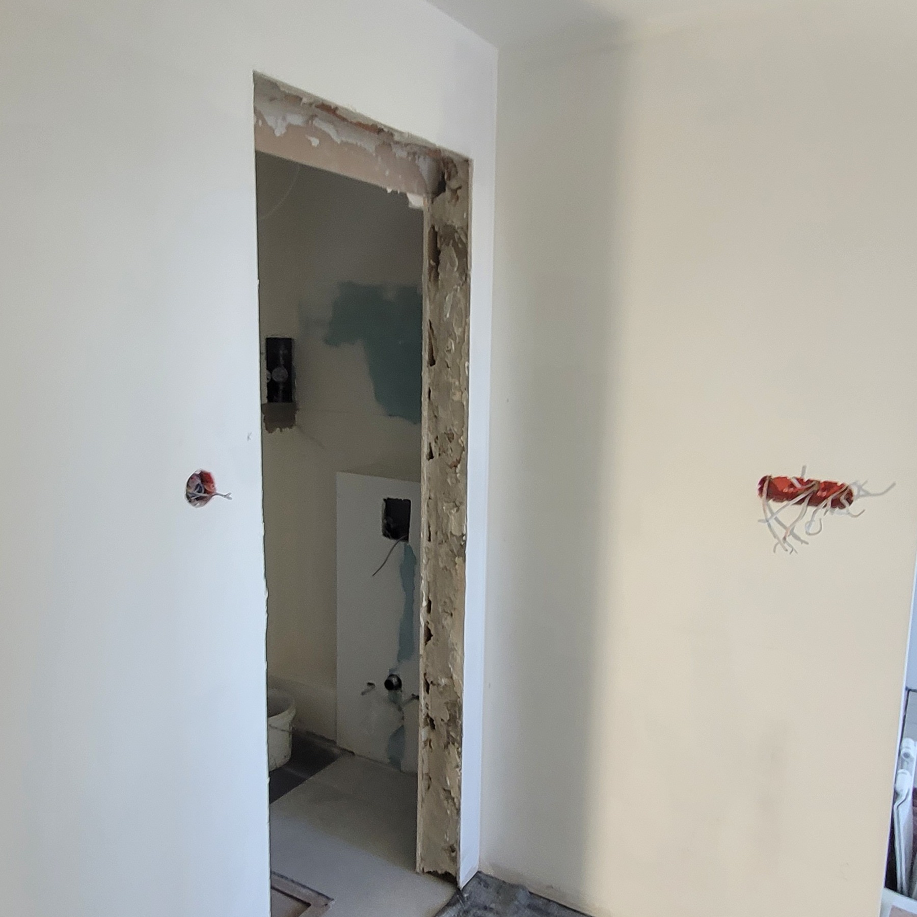 Wnętrze w trakcie remontu: widoczne ściany z otworami na instalacje elektryczne, surowe ościeże drzwiowe z pozostałościami zaprawy i fragment wyremontowanego pomieszczenia w tle.