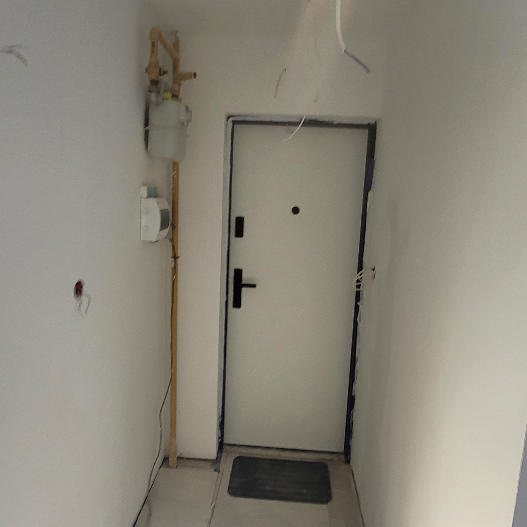 Korytarz w trakcie remontu z widoczną białą ścianą, licznikiem gazu, przewodami elektrycznymi wychodzącymi ze ściany oraz zabezpieczoną folią ościeżnicą drzwi wejściowych.