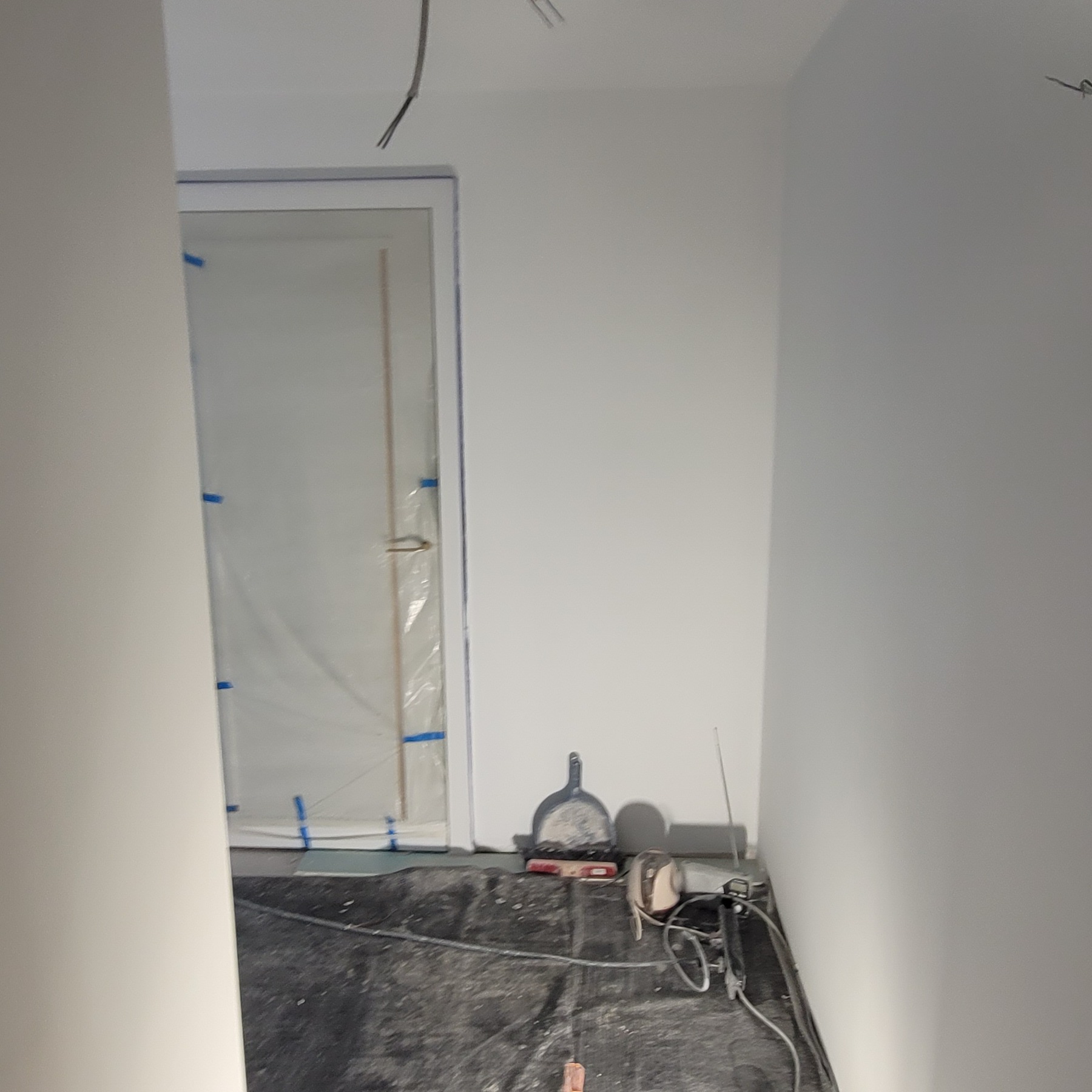 Przygotowanie do prac wykończeniowych: zabezpieczone folią i taśmą drzwi, narzędzia malarskie na podłodze pokrytej folią ochronną, ściany w trakcie szpachlowania.