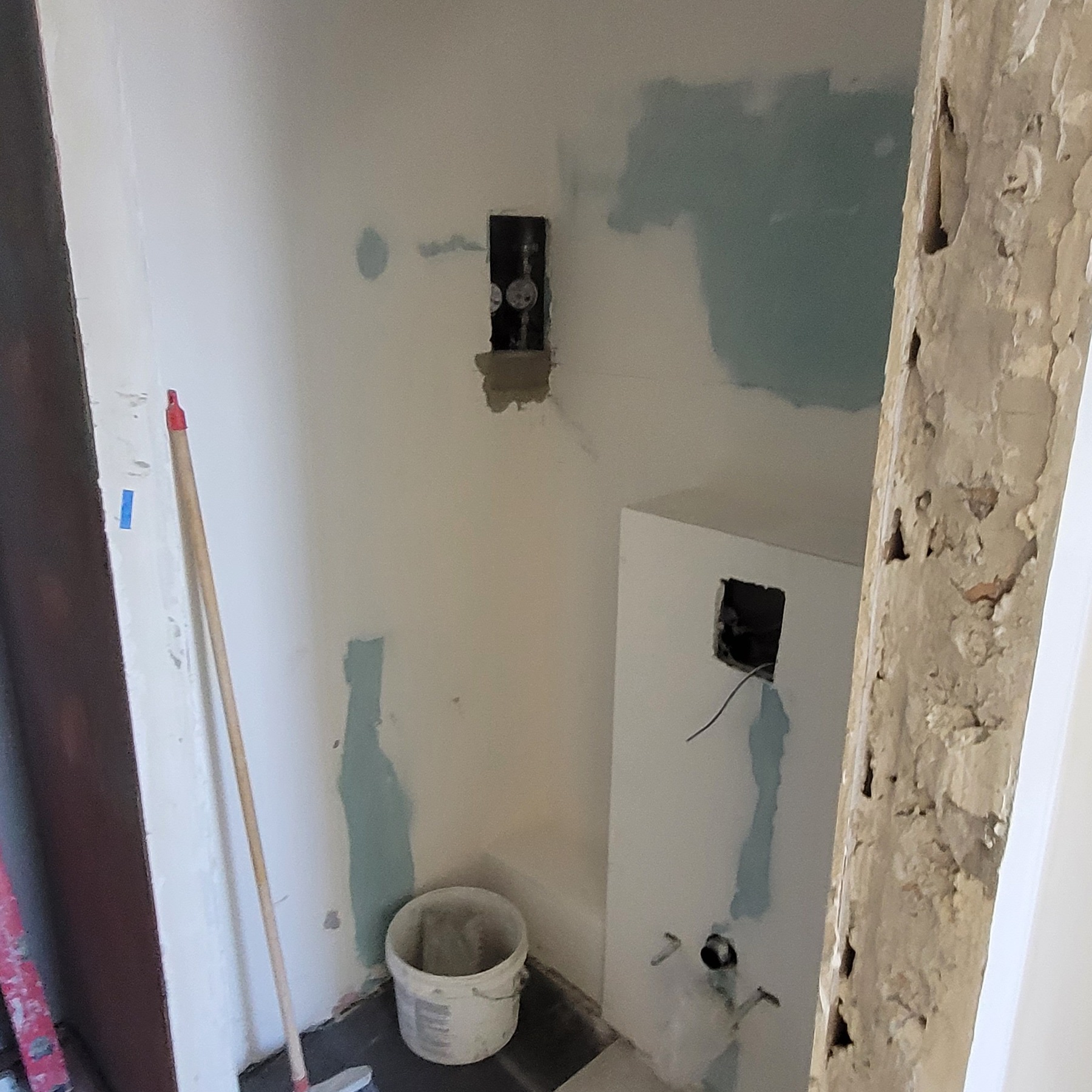 Wnętrze w trakcie remontu: przygotowane otwory pod instalacje elektryczne i wodne, ściany z widocznymi śladami po gładzi, wiadro i narzędzia na podłodze, fragment ościeżnicy z odpryskującą farbą.